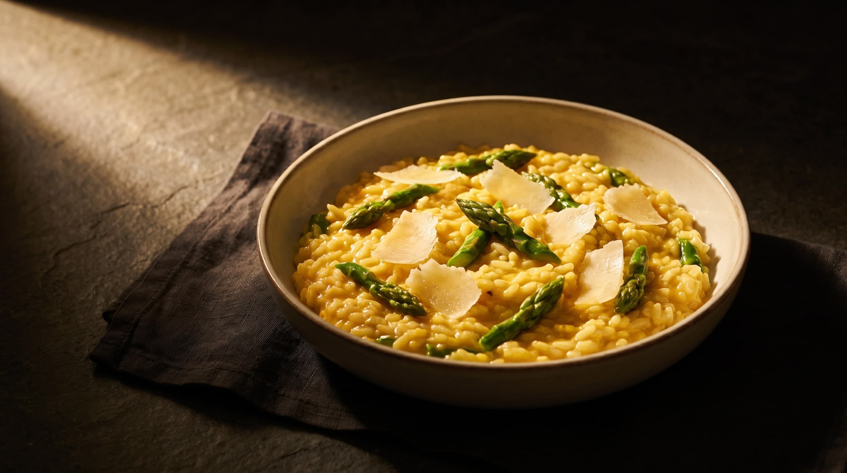 Safran-Risotto mit grünem Spargel und Parmesanspänen in heller Schale auf dunklem Stein