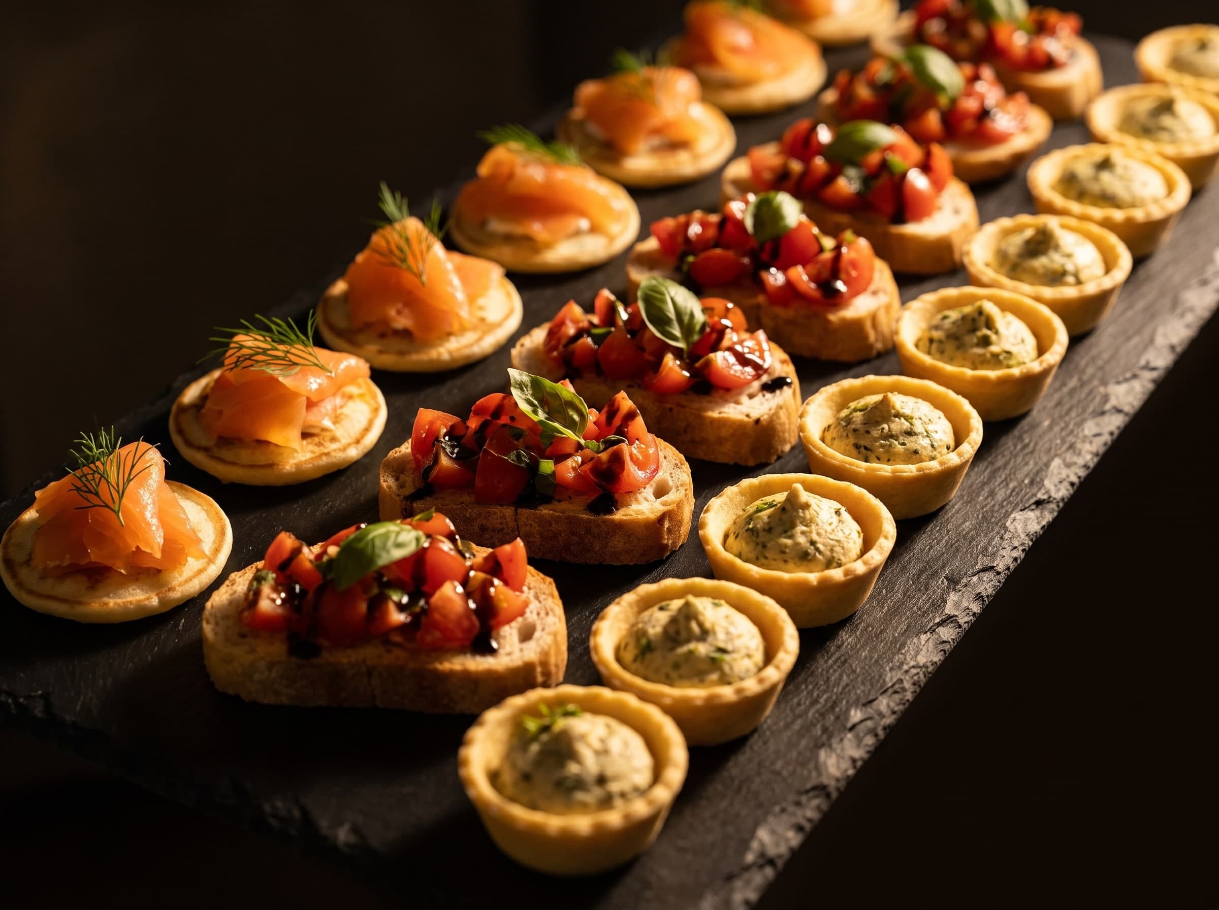 Dunkles Schieferbrett mit Reihen von Lachs-Blinis, Bruschetta und Kräutertartelettes